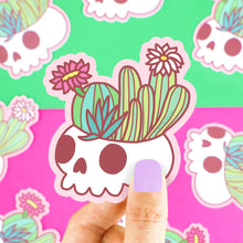 Load image into Gallery viewer, A hand holds a Turtle Soup Flowering Cactus Skull vinyl sticker, ideal for goth decor. Background is vibrant with plants.
