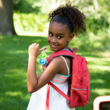 Load image into Gallery viewer, A smiling girl with curly hair holds Simpl Dimpl, Clear keychains and wears a red backpack in a sunny park. Brand: Fat Brain Toy Co.