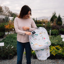 Load image into Gallery viewer, A woman in a garden holds a baby wrapped in the Milk Snob Cover Disney Enchanted Kingdoms, with plants filling the background.
