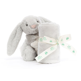 A Jellycat Bashful Silver Bunny Soother sits with a soft blanket, both tied with a white ribbon featuring green text and owl designs.