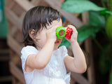 A toddler in a white dress uses PlanToys colorful cups as pretend Mushroom Kaleidoscope binoculars, surrounded by lush greenery.