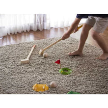 Load image into Gallery viewer, A child plays indoor mini golf with PlanToys sustainable Mini Golf Set, featuring wooden clubs and a flag hole setup on the carpet.