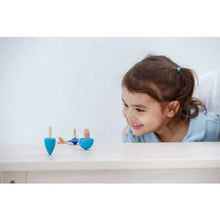 Load image into Gallery viewer, A girl in a blue shirt smiles as three PlanToys sustainably made Spinning Tops dance on the table.