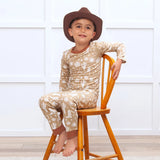 A boy in a cowboy hat smiles on a wooden chair in Tesa Babes Wild West Brown PJs, beige with white cactus patterns, eco-friendly.