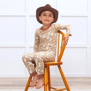 A boy in a cowboy hat smiles on a wooden chair in Tesa Babes Wild West Brown PJs, beige with white cactus patterns, eco-friendly.