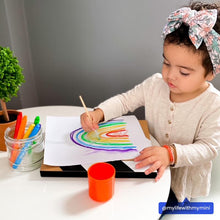 Load image into Gallery viewer, A toddler in a headband draws a vibrant rainbow at the table with OOLYs Smooth Stix Watercolor Gel Crayons; a plant peeks in.