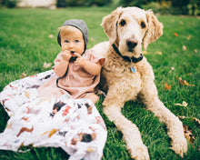 Load image into Gallery viewer, A baby in a pink dress and bonnet sits on grass next to a fluffy dog, with the Little Unicorn Woof Cotton Swaddle blanket nearby.