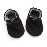 Cabooties Denim Black: Small black baby shoes with white stitching, ruffled edges, and flexible rubber soles on a white background.