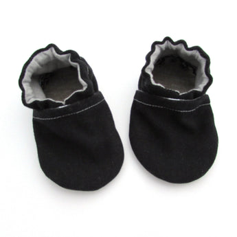Cabooties Denim Black: Small black baby shoes with white stitching, ruffled edges, and flexible rubber soles on a white background.