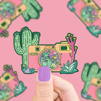 A person with lavender nail polish holds a Turtle Soup Vinyl Sticker, Desert Camera featuring a camera and cacti on a pink background.