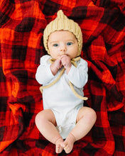 Load image into Gallery viewer, A baby with wide eyes lies under a Little Unicorn Cotton Swaddle, Red Plaid, wearing a knitted hat and white outfit.