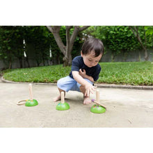 Load image into Gallery viewer, A child crouches to play PlanToys Meadow Ring Toss, aiming at pegs on concrete, honing fine motor skills with grass behind.