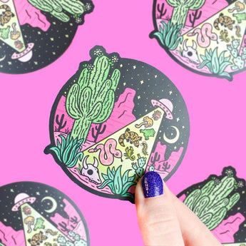 A hand holds Turtle Soups Vinyl Sticker, Desert Abduction—weatherproof with cacti, UFOs, and animals under a starry night.