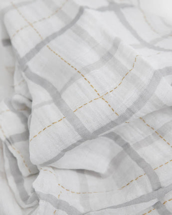 Little Unicorn Cotton Swaddle, Grey Plaid: white cotton muslin with a gray grid and yellow stitching, softly folded and slightly wrinkled.