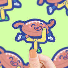 Load image into Gallery viewer, A Turtle Soup Vinyl Sticker shows a cartoon football character near a yellow goalpost on green, held by hand.