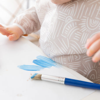 Close-up of a child wearing a Bumkins Long-Sleeved Art Smock: Wander, painting blue strokes on paper with a brush.