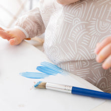 Load image into Gallery viewer, Close-up of a child wearing a Bumkins Long-Sleeved Art Smock: Wander, painting blue strokes on paper with a brush.