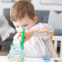 Load image into Gallery viewer, A child wearing a Bumkins Long-Sleeved Art Smock: Wander places a paintbrush into a jar, focused on creativity at a white table.