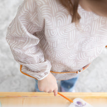 Load image into Gallery viewer, A child paints at an easel while wearing a Bumkins Long-Sleeved Art Smock: Wander, made of waterproof fabric.