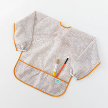 Load image into Gallery viewer, Bumkins Long-Sleeved Art Smock: Wander lays on a white background, holding two paintbrushes in the front pocket.