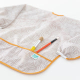 Bumkins Long-Sleeved Art Smock: Wander lays on a white background, with two paintbrushes in the front pocket.