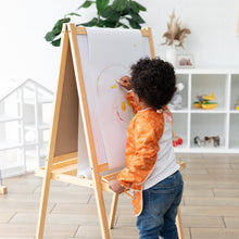 Load image into Gallery viewer, A child with curly hair draws on white easel paper in a bright room, wearing a Bumkins Long-Sleeved Art Smock: Grounded.