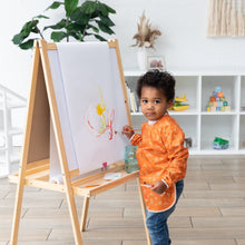 Load image into Gallery viewer, A child in a Bumkins Long-Sleeved Art Smock: Grounded paints at an easel, surrounded by shelves and plants in the bright room.