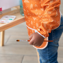 Load image into Gallery viewer, Close-up of a child wearing a Bumkins Long-Sleeved Art Smock: Grounded, holding a paintbrush next to an easel.