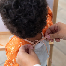 Load image into Gallery viewer, An adult fastens the neck tie of a Bumkins Long-Sleeved Art Smock: Grounded on a toddler.
