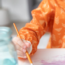 Load image into Gallery viewer, Close-up of a child in a Bumkins Long-Sleeved Art Smock: Grounded, painting beside a blurred glass jar.
