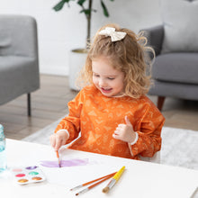 Load image into Gallery viewer, A smiling child with curly hair in a Bumkins Long-Sleeved Art Smock: Grounded paints on paper at a white table in a bright room.