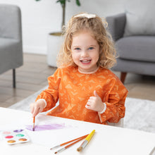 Load image into Gallery viewer, A young child with curly hair paints watercolors at a white table while wearing a Bumkins Long-Sleeved Art Smock: Grounded.