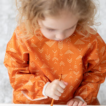 Load image into Gallery viewer, A child wears a Bumkins Long-Sleeved Art Smock: Grounded, paintbrush in hand, focused on painting.