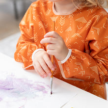 Load image into Gallery viewer, A child in a Bumkins Long-Sleeved Art Smock: Grounded paints with purple and gray paints on paper.