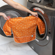 Load image into Gallery viewer, A person holds a Bumkins Long-Sleeved Art Smock: Grounded in front of an open front-loading washing machine.