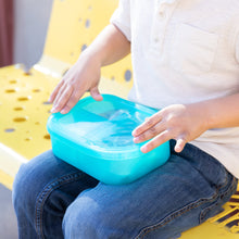 Load image into Gallery viewer, A child sits on a yellow bench, holding a Bumkins Silicone Bento Box 3 Section: Blue Jelly on their lap.