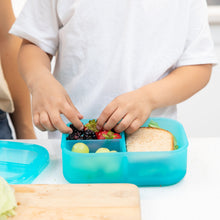 Load image into Gallery viewer, A child packs fruit and a sandwich into a Bumkins Silicone Bento Box 3 Section: Blue Jelly on a kitchen counter.