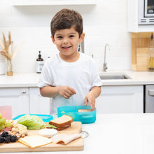 Load image into Gallery viewer, A smiling child makes a sandwich in a bright kitchen, surrounded by ingredients and a Bumkins Silicone Bento Box 3 Section: Blue Jelly.