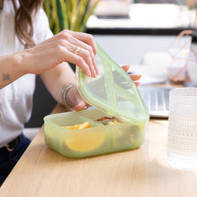 Load image into Gallery viewer, An adult opens a Bumkins Silicone Bento Box 3 Section: Green Jelly filled with snacks on a desk next to an ornate glass cup.
