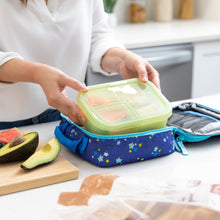 Load image into Gallery viewer, An adult places a Bumkins Silicone Bento Box 3 Section: Green Jelly into an open blue lunch bag next to avocado slices on a kitchen counter.