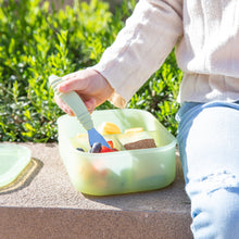 Load image into Gallery viewer, A child uses Bumkins Spoon + Fork: Sage to eat from a green bento box on a concrete bench by greenery.