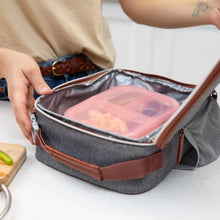 Load image into Gallery viewer, A person puts a Bumkins Silicone Bento Box 3 Section: Pink Jelly into a gray insulated lunch bag on a kitchen counter.
