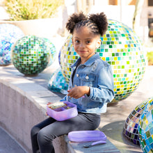 Load image into Gallery viewer, A curly-haired girl sits on a bench with a Bumkins Silicone Bento Box 3 Section: Purple Jelly, against colorful mosaic spheres.