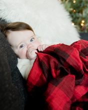 Load image into Gallery viewer, A blue-eyed baby lies on a couch with Little Unicorns red plaid Cotton Swaddle, a white pillow, and a Christmas tree behind.
