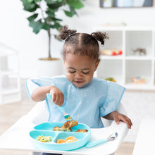 Load image into Gallery viewer, A young curly-haired girl sits in a high chair, wearing a Bumkins Ruffle Apron Bib: Cinderella while eating from a blue plate with utensils.