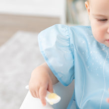 Load image into Gallery viewer, Close-up of a baby in a Bumkins Ruffle Apron Bib: Cinderella holding a spoon, with focus on the ruffle sleeve.
