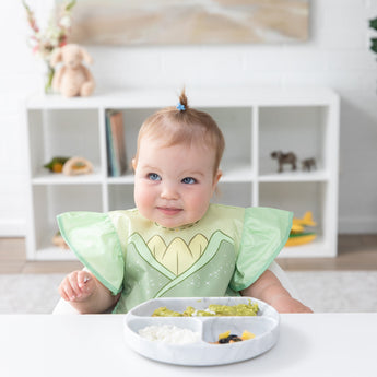 A baby in a Bumkins Ruffle Apron Bib: Tiana sits at a white table with a gray plate of food, looking off to the side with a soft smile.