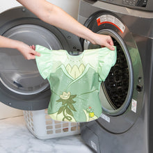 Load image into Gallery viewer, A person holds a Bumkins Ruffle Apron Bib: Tiana in front of an open front-loading washing machine.