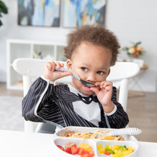 Load image into Gallery viewer, A toddler in Bumkins Sleeved Bib: Jack Skellington playfully holds a spoon to their mouth while sitting at a table with a plate of food.