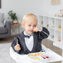Load image into Gallery viewer, A toddler wearing Bumkins Sleeved Bib: Jack Skellington enjoys food from a divided tray, sitting in a high chair with a cozy living room view.